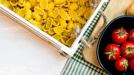 Top view of pasta in a wooden crate and fresh ripe cherry tomatoes in a black bowl on a rustic white wooden table. Ingredients and food conceptの写真素材