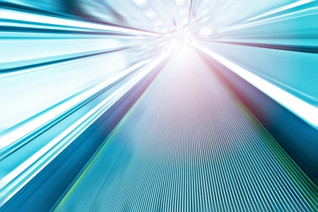 Perspective wide angle view of modern light blue illuminated and spacious  high-speed moving escalator with fast blurred trail of handrail in vanishing traffic motionの写真素材