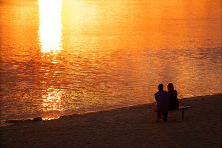 A man and a woman sit on a bench on the shore and admire the sunset, the light of the sun is reflected on the waterの写真素材