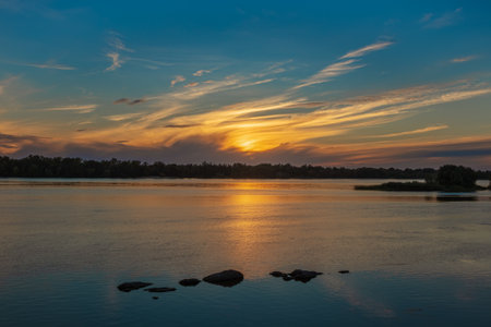 Beautiful sunset over the Dnieper River. Yellow cirrus clouds over the forest. Sun light reflection and stones in waterの写真素材