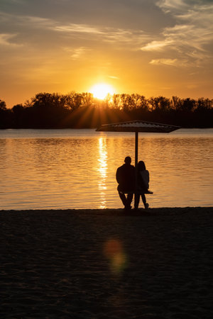A couple in love admires the setting Sun over the river and forest. Silhouette of a man and a woman on the beach. Romance and beauty of natureの写真素材