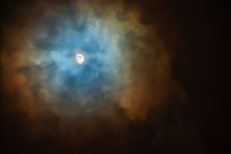 The Moon in the sky on a cloudy night is surrounded by colored clouds. Corona - in meteorology plural coronae is an optical phenomenon produced by the diffraction of moonlight by water droplets or tiny ice crystals of a cloud.の写真素材