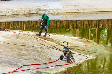 Kharkiv city, Ukraine - April 12, 2024: A man cleans the bottom of an artificial pond with high-pressure washer nozzle from mud and sludge. Spring work on caring for a pondのeditorial素材
