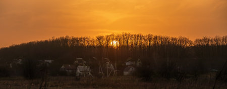 Panoramic view of the landscape with the setting Sun behind the trees on the horizon. Yellow-orange sunset over a forest and meadow in the countrysideの写真素材