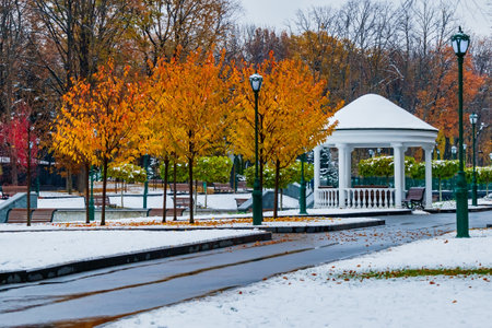 Autumn park covered with first snow. Alley with trees in yellow-orange leaves and white gazebo in snowy landscape of city parkの写真素材