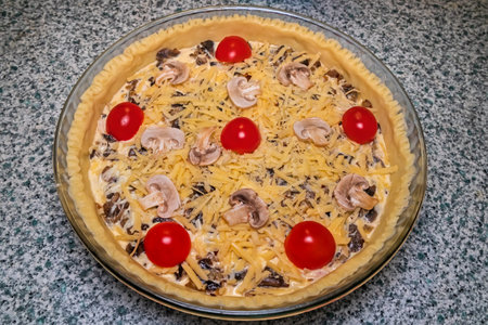 Quiche tart in the process of cooking before baking. Sliced ââmushrooms, yellow grated cheese and red cherry tomatoes on shortcrust pastry. Glass plate on the tableの写真素材
