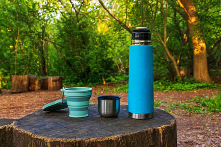 Thermos with tea or coffee and silicone folding cup on a wooden stump. Drinkware for a picnic or a walk. Drink outdoors in the forestの写真素材