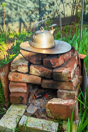 Stainless steel kettle on homemade outdoor oven made of bricks. Oven on grassy area of garden. Rural old way of boiling waterの写真素材
