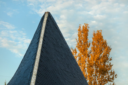 View of a peaked roof, blue sky with clouds and a tree with yellow autumn leavesの写真素材