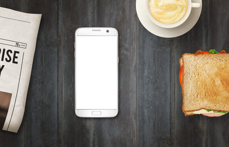 Smart phone with isolated display for mockup on table. Newspapers, coffee and sandwich on table. Top...