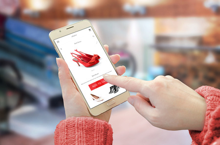 Shopping web site app on smart phone. Woman holding mobile device and buy red shoes. City shopping center in background.の写真素材