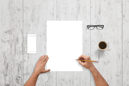 Man write on empty paper on white wooden desk. White smart phone with isolated screen for mockup. Glasses and cup of coffee beside.の写真素材