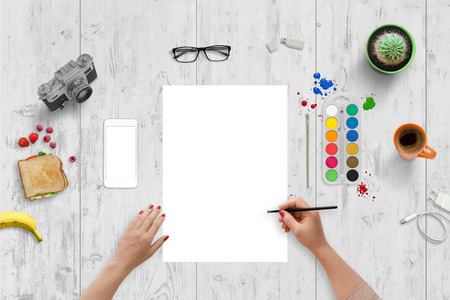 Woman painting. Empty clean paper for art presentation. Top view of white wooden desk. Water colors, mobile phone, classic camera, sandwich, coffee, glasses, plant, cactus, charger beside.の写真素材