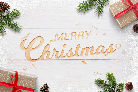 Merry Christmas text hand carved in white woodej surface surrounded with Christmas gifts, fir branches and pinecones. Snowflakes beside. Top view.の写真素材
