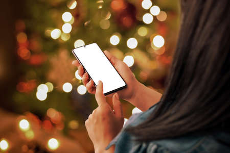 Christmas phone mockup in woman hand. Sending Christmas greetings by phone concept. Bokeh lights of decorated Christmas tree in backgroundの写真素材