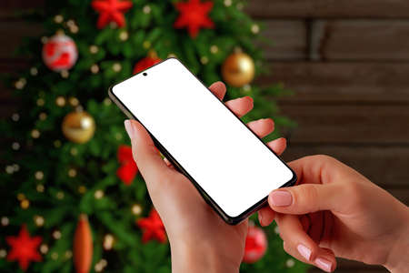 Christmas phone mockup concept. Smart phone in woman hands front off Christmas tree. Isolated display for app promotionの写真素材