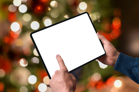 Tablet in the hands with the lights of the Christmas tree behind. Isolated display for design promotion. Season online shopping conceptの写真素材