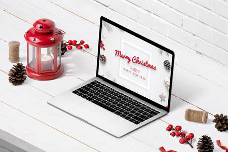 Laptop with Merry Christmas and Happy New Year greeting text on display. Isometric composition on white wooden desk with Christmas decorationsの写真素材