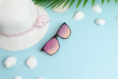 Woman hat and glasses on blue surface surrounded by shells and palm leaves. Summer vacation...