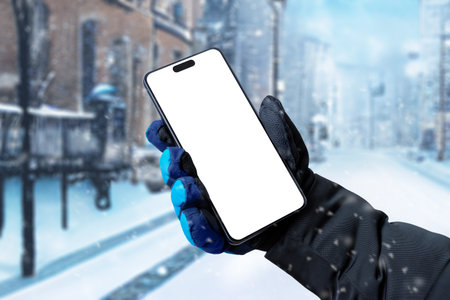 male hand in a black glove holds a smartphone with a white screen on the background of a snowy city streetの写真素材
