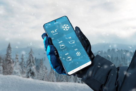 Smartphone with smart home application on the screen in the hands of a woman in black gloves against the background of a snowy mountain landscapeの写真素材