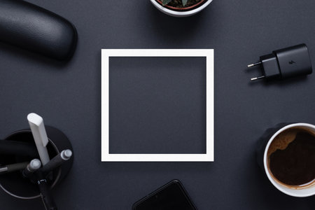 White square paper frame on black office desk surrounded by coffee mug, charger, plant, pens and phone. Copy space in the middle. Top view flat layの写真素材