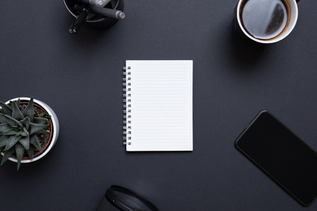 Blank notepad on black desk surrounded by coffee mug, phone, plant, pens and camera lens. Concept business flat lay compositionの写真素材