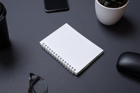 Blank notepad on black work desk witc copy space. Surounded by office supplies, phone, glasses, coffe mug, plant. Low light business compositionの写真素材