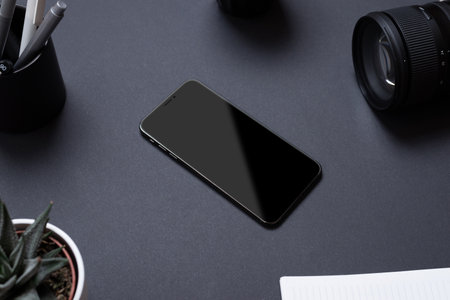 Black office desk table with blank smartphone surounded by camera lens, pens, plant and notepad. Business compositionの写真素材