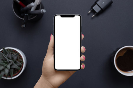 Phone in woman hand with isolated display for mockup. Business, office mockup for app or web page presentation. Office desk with coffee mug, plant, pens, charger in backgroundの写真素材