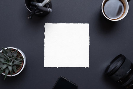 Blank paper frame on black office desk surrounded by phone, camera, coffee mug, plant and pens. Top view, flat lay composition for copy presentationの写真素材