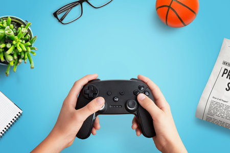 Top view flat lay composition of a player holding a gamepad on a desk with a ball, newspaper,...