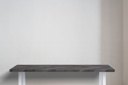 Industrial-style black desk with wooden texture and white legs, set against a white wall with soft window shadows, ideal for modern office setups and tech product displayの写真素材