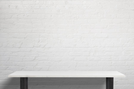 Minimalist workspace featuring a sleek white table with black legs, bright lighting, and a white brick background, perfect for creative work environmentsの写真素材