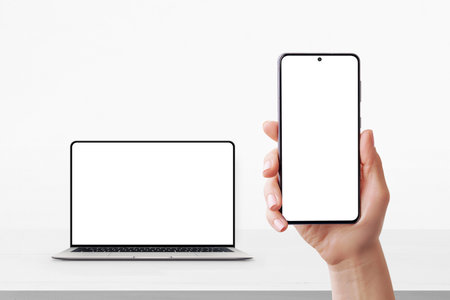 Smartphone in hand with blank screen and laptop in background on desk, both ready for UI or branding mockupの写真素材