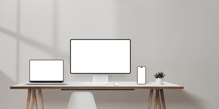 Bright office desk with laptop, computer monitor, and smartphone mockups, blank wall space for promotional copyの写真素材