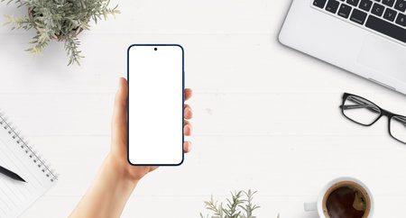 Top view of smartphone in female hand over white desk with laptop, coffee, notebook, and workspace items. Ideal for app mockup or UI displayの写真素材
