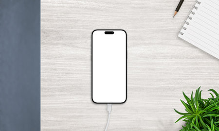 Top view of smartphone on charger with blank screen, placed on desk with notebook and plant. Ideal for productivity and app mockupsの写真素材