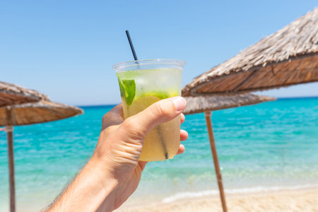 Hand holding refreshing cocktail with lime and mint against tropical beach backdrop, perfect for summer and vacation conceptsの写真素材