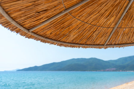 Straw beach umbrella shading view of blue sea and distant mountains on a sunny summer dayの写真素材