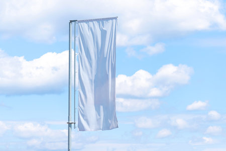 Single tall vertical white banner on pole fluttering in wind with cloudy blue sky background, outdoor advertising spaceの写真素材