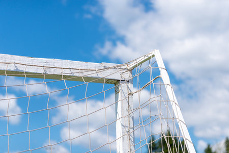 Close-up of worn football goalpost corner with net against blue sky and white clouds.の写真素材
