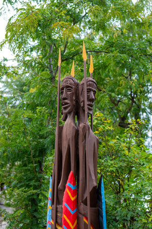 Tall wooden tribal statue with colorful shields and spears, representing indigenous warrior figures, surrounded by lush green trees outdoors.の写真素材