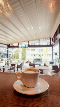 Vertical shot of coffee cup on wooden table inside bright modern cafe, relaxing atmosphere ideal for hospitality, design, and lifestyle themesの写真素材