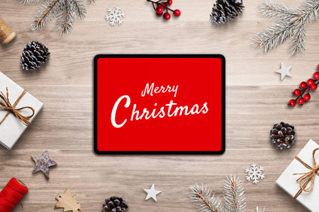 Tablet on wooden table with red âMerry Christmasâ screen surrounded by pine cones, stars, branches and gifts, top view festive flat layの写真素材