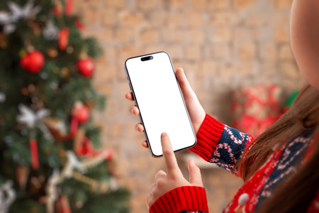 Hand holding smartphone with blank screen in cozy Christmas setting, ideal for app mockups, holiday promotions, mobile UI presentations and festive marketing visualsの写真素材