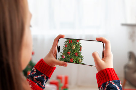 Woman photographing a decorated Christmas tree with smartphone, capturing festive ornaments and cozy holiday atmosphere in a bright winter home settingの写真素材