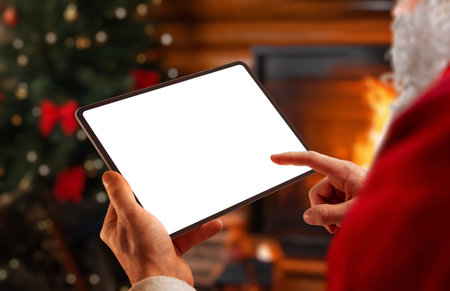 Santa holding tablet with blank screen in cozy Christmas interior, ideal for holiday mockups, winter marketing, festive apps and seasonal digital designsの写真素材
