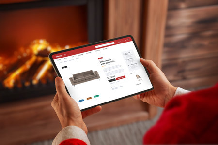 Santa browsing online furniture store on tablet near fireplace, ideal for ecommerce themes, holiday shopping concepts, festive marketing and winter retail promotionsの写真素材