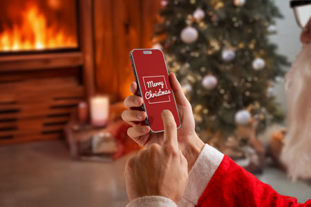 Santa using smartphone with merry christmas screen near fireplace and decorated tree, perfect for festive marketing, holiday greetings, winter campaigns and seasonal digital contentの写真素材